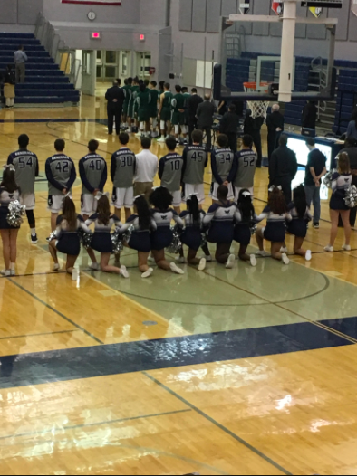 Nine cheerleaders kneel during the national anthem, while the rest of the team and the basketball team stands